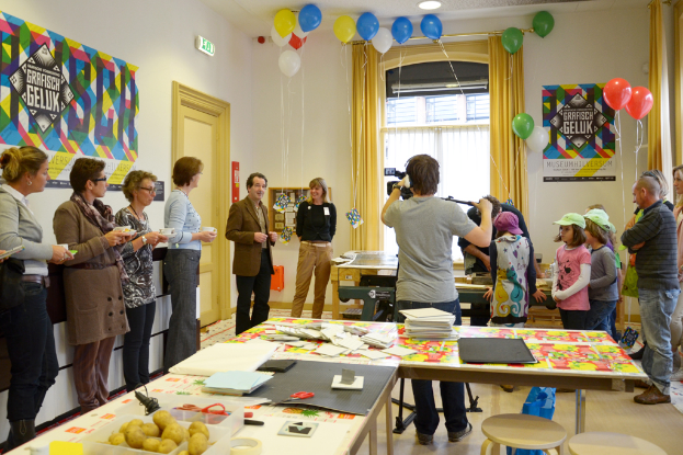 Ein voller Raum mit feiernden Menschen, Tischen mit verschiedenen Gegenständen wie Büchern und Kartoffeln, ein Mann mit einer Kamera auf einem Stativ, Menschen mit Tassen und Kinder mit Mützen in der Ecke, mit einem Fenster und Vorhängen im Hintergrund.