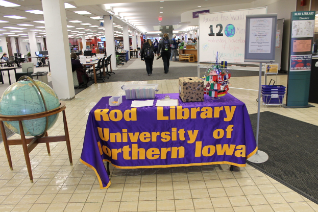 Eine Ausstellung mit einem Tisch, auf dem verschiedene Landesflaggen, eine Box, Papiere und ein Banner stehen, eine Kugel zur Linken, ein Anschlagbrett zur Rechten mit einigen Menschen in der Nähe und mehreren Tischen und Stühlen zur Linken.