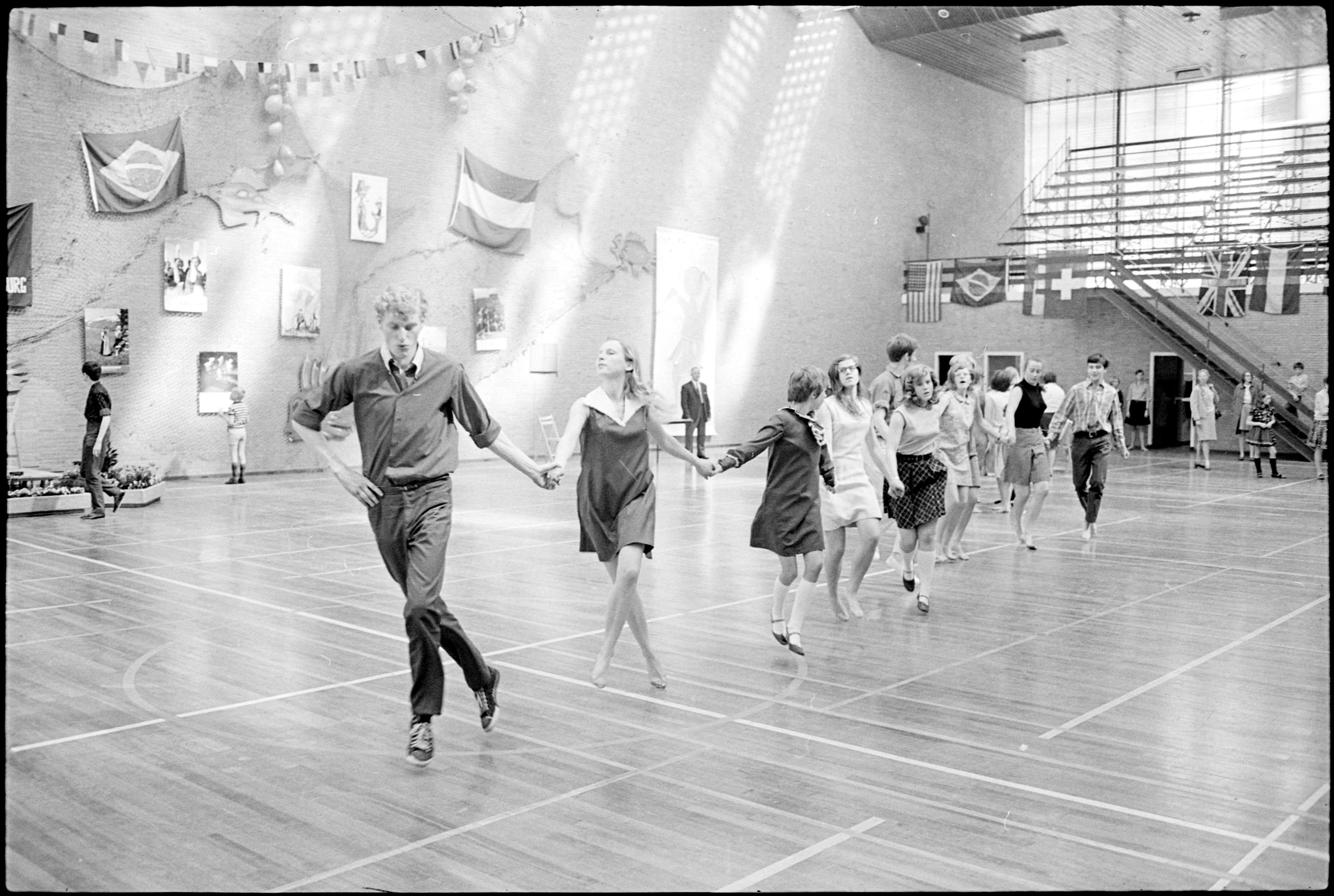 Schwarzes und weißes Foto von Menschen, die in einem Turnsaal tanzen, mit Foto Rahmen und Flaggen an der Wand, Stufen auf der rechten Seite.