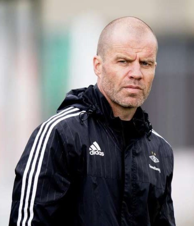 Ein kahlköpfiger Trainer in einer blauen Jacke mit weißen Streifen und einem weißen Kragen steht auf einem Fußballfeld, die Hände gefaltet, mit einem ernsten Gesichtsausdruck und einem unscharfen Hintergrund.