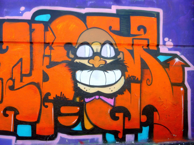 Graffiti-Gemälde eines Cartoon-Charakters mit großen Augen, einem breiten Grinsen und einem verschmitzten Gesichtsausdruck an der Seite eines Gebäudes, mit leuchtenden Farben und einer mutigen Begleittext.