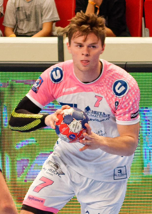 Ein Mann in einem pinken Hemd, der einen Fußball hält, mit der Hand eines anderen Menschen links daneben und mehreren Personen im Hintergrund auf Stühlen sitzend.