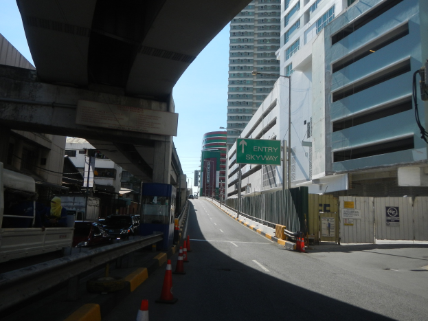 Stadtstraße mit Verkehrskegeln am Straßenrand, fahrende Fahrzeuge, eine Brücke auf der linken Seite, Gebäude mit Schildern auf beiden Seiten, Laternenpfähle und ein Himmel im Hintergrund.