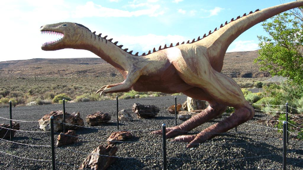 Eine Dinosaurierstatue steht mit einigen Gegenständen auf dem Boden und Bäumen und dem Himmel im Hintergrund.