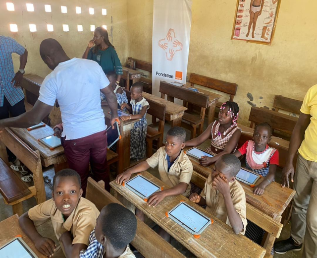 Kinder an Tischen mit Tablets in einem Klassenzimmer mit Erwachsenen in der Nähe, ein Banner und ein Plakat an der Wand.