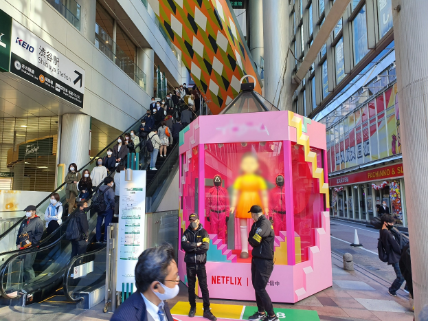 Eine Gruppe von Menschen steht vor einem Gebäude mit Glaswänden und Texttafeln, einige tragen Masken, während eine Rolltreppe mit Menschen daneben fährt; ein pinker Kasten in der Mitte zeigt Schaufensterpuppen und trägt "Netflix's new Netflix booth in Tokyo."