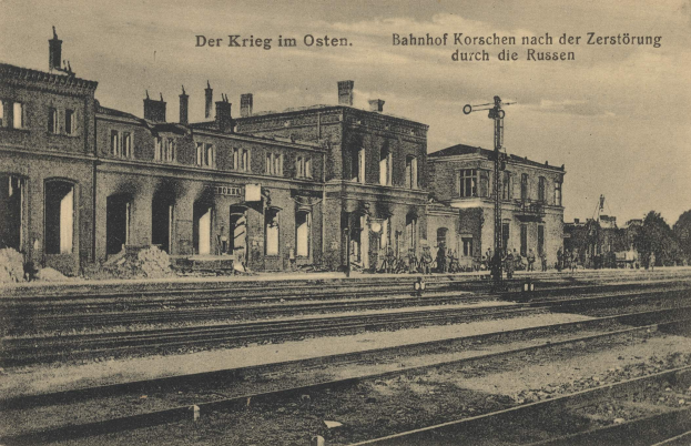 Schwarz-weißes Foto des Bahnhofs Korschen in Ostern, Deutschland, mit Bahnschienen, Gebäuden, Strommasten, Menschen, Bäumen und Himmel.