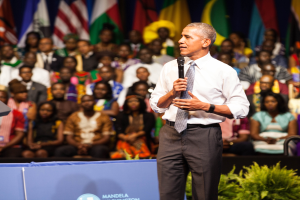 Präsident Obama spricht auf einer Wahlveranstaltung, hält ein Mikrofon, mit einer Menschenmenge, einige sitzen, einige stehen, und einer Pflanze im Vordergrund, Fahnen und eine Tafel im Hintergrund.