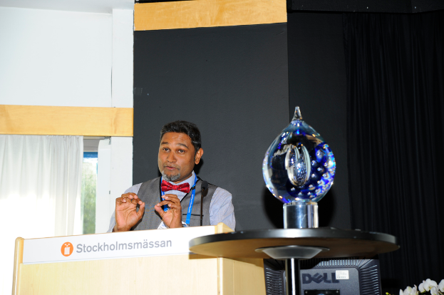 Ein Mann steht neben einem Rednerpult und spricht mit einer Wand dahinter.