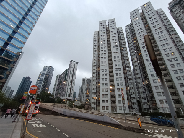 Eine Stadtstraße mit hohen Gebäuden, Fahrzeugen, Fußgängern, Schildern, Laternen, Bäumen und einem bewölkten Himmel, mit einem Wohnhaus im Vordergrund.