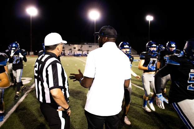 Fußballtrainer in Mütze spricht mit seinem Team auf einem nächtlichen Feld, mit Spielern in Helmen, Laternenmasten und dunklem Himmel im Hintergrund.