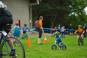 Kinder fahren Fahrräder auf einem Weg mit Kegelabsperrungen, während vier Personen in der Nähe stehen; eine Wand, Bäume und der Himmel sind im Hintergrund zu sehen.