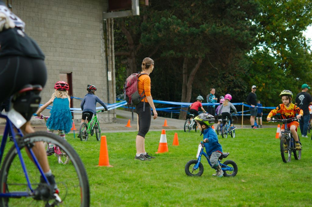 Kinder fahren Fahrräder auf einem Weg mit Kegelabsperrungen, während vier Personen in der Nähe stehen; eine Wand, Bäume und der Himmel sind im Hintergrund zu sehen.