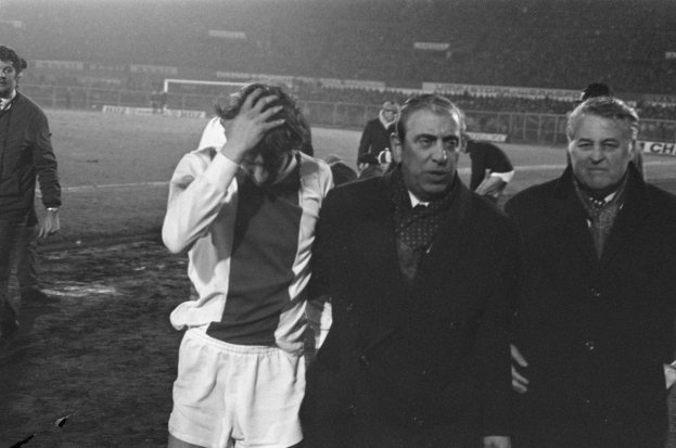Eine Gruppe von Männern in Not steht zusammen auf einem Fussballfeld, mit einem Stadion im Hintergrund, auf einem Schwarz-Weiss-Bild.