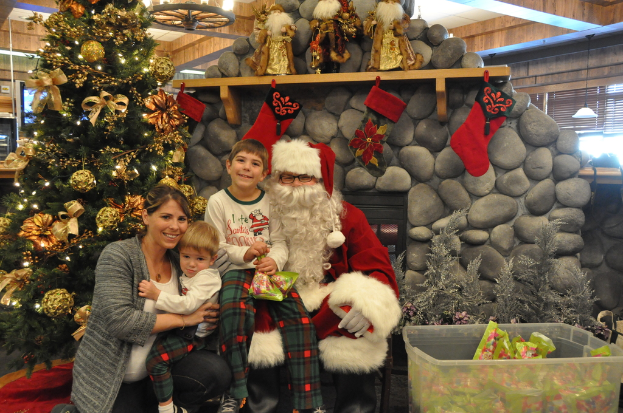 Eine Frau, Kinder und ein Mann in Santa-Klaus-Kostüm mit Brille und Handschuhen stehen in der Nähe eines geschmückten Weihnachtsbaums, einer Wand mit Felsen, Spielzeugen, Dekorationen und einem Korb mit Gegenständen, mit Lichtern im Hintergrund.