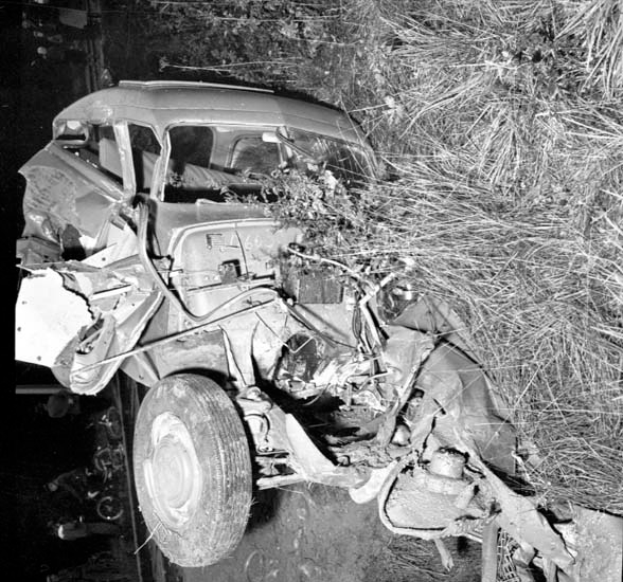 Ein Schwarz-Weiß-Foto eines schwer beschädigten Autos am Straßenrand mit einer eingedrückten Front, zerschlagenen Fenstern, einem Baum im Hintergrund und verstreuten Gegenständen auf der Szene.