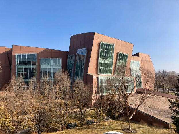Großes Gebäude mit vielen Fenstern, umgeben von Bäumen, Bänken, Gras und einem Laternenmast, mit anderen Gebäuden und einem klaren blauen Himmel im Hintergrund, als der neue Campus der University of Illinois at Chicago identifiziert.