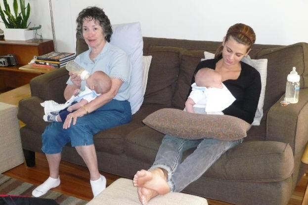 Zwei Frauen sitzen auf einem Sofa und halten Babys, mit Kissen auf dem Sofa, einer weißen Wand im Hintergrund und einem Schreibtisch mit Büchern und einer Zimmerpflanze links.