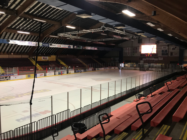 Eine große Indoor-Hockeyhalle mit Sitzplätzen, einem Zaun, einem Netz, Bannern, einem Bildschirm und Deckenlampen, umgeben von verschiedenen Gegenständen.