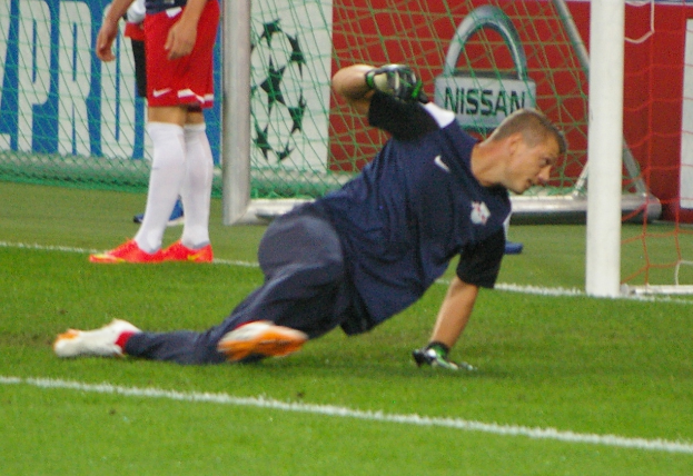 Ein Füßballspieler in Handschuhen taucht ab, um einen Ball vor einem Tor zu fangen, mit zwei Personen, einem Netz und einer Banner mit Text im Hintergrund.