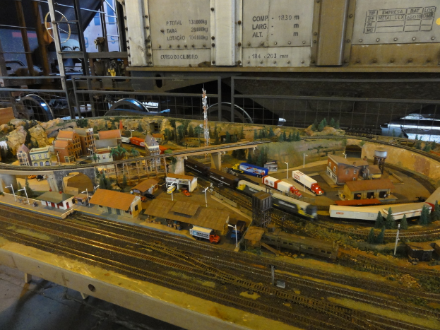 Eine Miniaturlandschaft auf einem Tisch mit einer Schienenstrecke, einem Zug, Fahrzeugen, Gebäuden, einer Brücke, einem Turm und Bäumen.