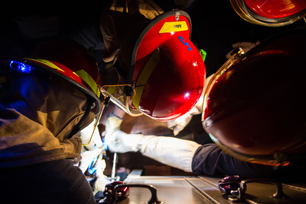 Feuerwehrleute in Helmen und Handschuhen arbeiten an einem Feuerwehrauto in einem schwach beleuchteten Bereich mit einem Tisch voller Notfallversorgungsmittel auf der rechten Seite.