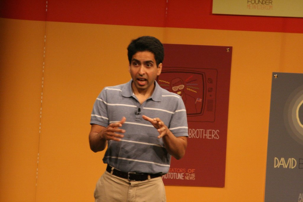 Ein Mann steht auf einer BÃ¼hne und spricht mit einer Wand voller Plakate dahinter.