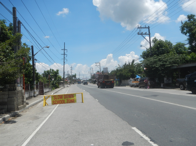 Eine Straße mit fahrenden Fahrzeugen, ein "Slow Down School Zone"-Schild, Strommasten mit Drähten, Bäume, Laternenpfähle und ein bewölkter Himmel im Hintergrund.