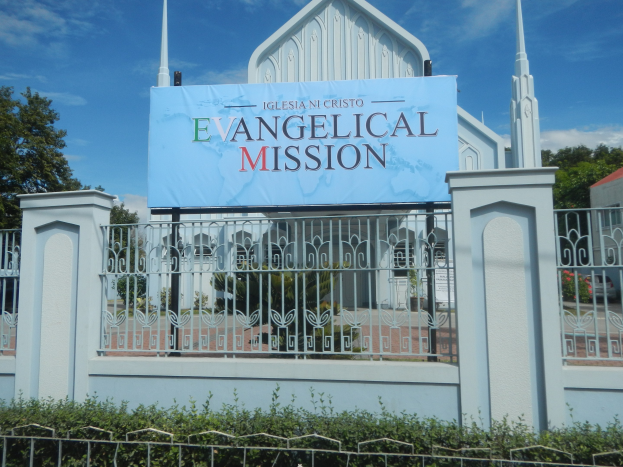 Kirche mit Metallzaun, einem Schild mit der Aufschrift "Evangelische Mission", umgeben von Bäumen, einem Haus mit Dach und Pflanzen unter einem bewölkten Himmel.
