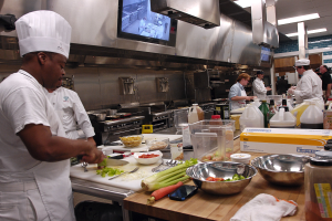 Eine Gruppe von Köchen in einer Küche, die Essen auf einem Schneidbrett zubereiten, mit einem Koch im Vordergrund in einem weißen Kleid und einer Mütze, der ein Messer hält und von Utensilien, Schüsseln, Gemüse, einem Mobiltelefon, Flaschen und anderen Gegenständen umgeben ist, mit einem Fernseher und Deckenlampen im Hintergrund.
