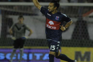 Ein Fußballspieler feiert nach einem Tor auf dem Boden, mit einer jubelnden Menge und einer Person im Hintergrund und einer Texttafel dahinter, alles vor einem leicht unscharfen Hintergrund.