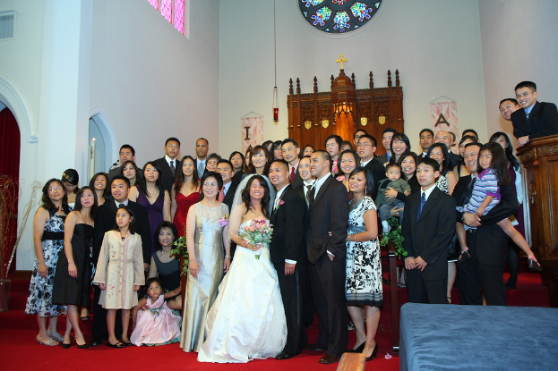 Eine Gruppe von Menschen auf einem roten Teppich, mit einer Frau in einem weißen Kleid und Schleier in der Mitte, einer Bühne mit blauer Stoff rechts, einem Podium in der rechten oberen Ecke, hölzernen Strukturen und einer Wand mit Türen und Fenstern links.