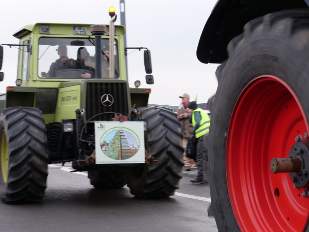 Ein grüner Traktor mit einem roten Rad fährt auf einer Straße voller Menschen in Jacken und Mützen, mit Bäumen und einem klaren blauen Himmel im Hintergrund.