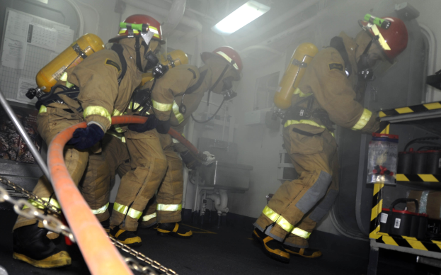 Feuerwehrleute in Schutzausrüstung und Helmen arbeiten an einem Löschfahrzeug, mit einem Gestell von Gegenständen und Hintergrundrohren und Lichtern.