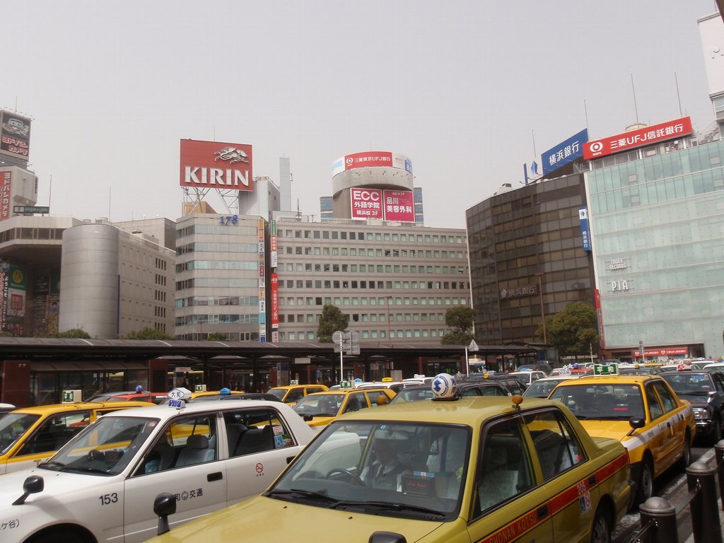 Eine Stadtansicht mit Fahrzeugen auf der Straße, Gebäuden, Bäumen, Pfählen mit Schildern, Text auf den Schildern, einer Bahn im Hintergrund und einem Geländer unten rechts.