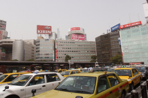 Eine Stadtansicht mit Fahrzeugen auf der Straße, Gebäuden, Bäumen, Pfählen mit Schildern, Text auf den Schildern, einer Bahn im Hintergrund und einem Geländer unten rechts.