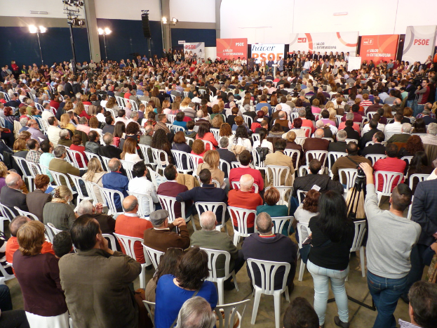 Ein volles Stadion mit vielen sitzenden und stehenden Menschen, eine Bühne mit zwei Rednern auf einem Podium und mehrere Banner und Lichter in der Nähe.