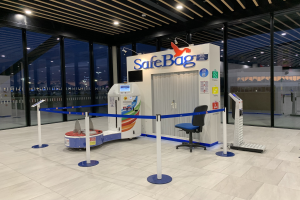 Sicherer Koffer-Eincheckschalter am Flughafen mit Stuhl, Absperrpoller, Maschinen, Bildschirm, bedruckter Tafel, Glaswänden, Deckenleuchten und sichtbarem Boden.