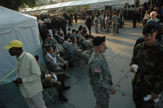 Eine große Menschenansammlung im Freien in militärischer Kleidung, wobei einige in der Mitte auf Stühlen sitzen und ein Baum oben im Bild zu sehen ist.