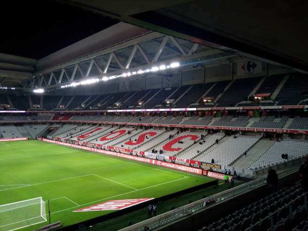 Großes Stadion mit einem Fußballfeld, das von grünem Gras bedeckt ist, einem Netz auf der linken Seite, einigen Menschen auf den Tribünen und hellen Deckenlampen, die die Szene vor einem dunklen Hintergrund beleuchten.