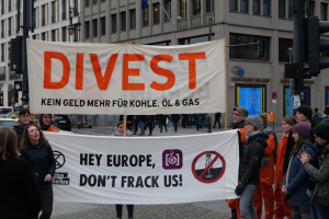 Gruppe von Menschen mit einem Banner mit der Aufschrift "Divest Hey Europe, Don't Frack Us" auf einer Straße mit Gebäuden, Fahrzeugen und Bäumen im Hintergrund unter einem klaren blauen Himmel.