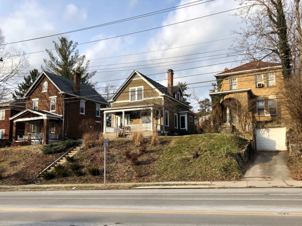 Reihe von Häusern mit Fenstern, Türen, Säulen, Geländern, Treppen, Pflanzen und einem Pfahl mit einer Tafel, vor einem bewölkten Himmel.