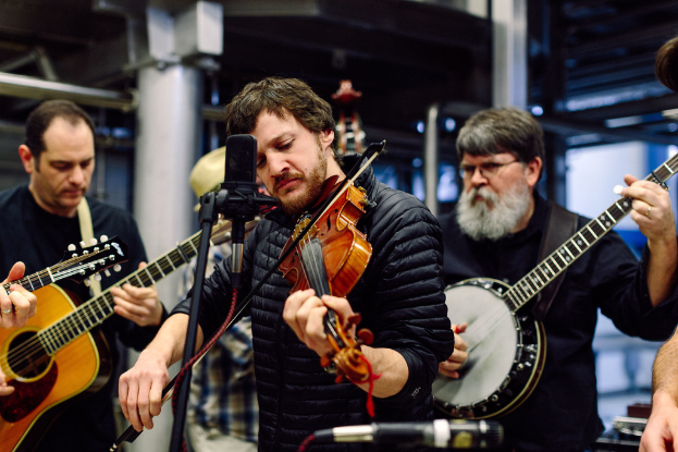 Eine Gruppe von Musikern in schwarzer Kleidung, die Instrumente spielen, mit einem Geiger vor einem Mikrofon und einem Gebäude im Hintergrund.