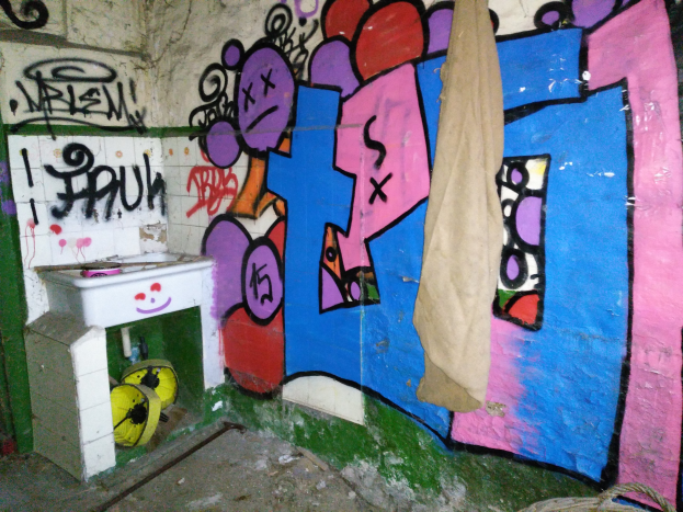 Ein mit Graffiti bedecktes Badezimmer mit einem Waschbecken in der Ecke, ein Tuch über dem Waschbecken und verstreute Gegenstände auf dem Boden.