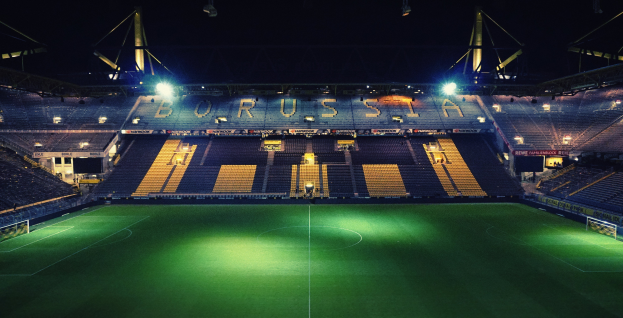 Ein Fubballfeld bei Nacht, beleuchtet von hellen Stadionlichtern, mit einigen Menschen auf den R├Ąngen und saftig gr├╝nem Rasen.