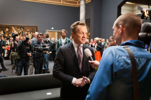 Ein Mann im Anzug und mit Krawatte spricht mit einem Reporter und hält ein Mikrofon in der Hand, während er von einer Menschenmenge umgeben ist, einige halten Kameras, im Hintergrund ein Sofa und eine Wand mit einem Fotorahmen und Lampen.