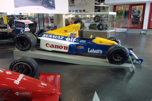 Eine Museumsausstellung mit verschiedenen Rennwagen, darunter ein Renault F1-Auto, das von Besuchern betrachtet wird, ein Textschild an der Wand, Deckenleuchten und Glastüren auf der rechten Seite.