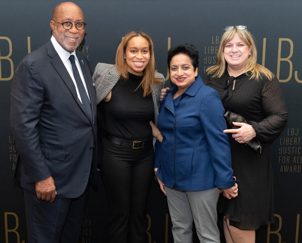 Eine Gruppe von vier Menschen auf einem roten Teppich, die lächeln und für ein Foto posieren, mit einer Tafel im Hintergrund, auf der 'Liberty Justice for All Awards' steht.