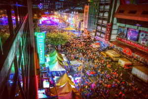 Eine bunte Karnevalszene mit einer großen Menschenmenge auf der Straße, umgeben von Zelten, Geschäften, Gebäuden und Bannern, mit zahlreichen Lichtern im Hintergrund.