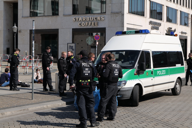 Eine Gruppe von Polizeibeamten steht vor einem Starbucks Café, rechts daneben ein Van und links ein paar Menschen; im Hintergrund ist ein Gebäude mit Fenstern, ein Schild, ein Laternenmast und ein Zaun zu sehen.